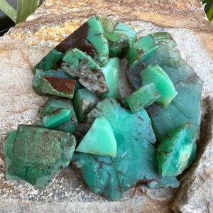 May include: A collection of green and brown chrysoprase gemstones, some with a rough, natural texture, others with a smooth, polished surface. The stones are arranged in a pile on a light brown, textured surface.