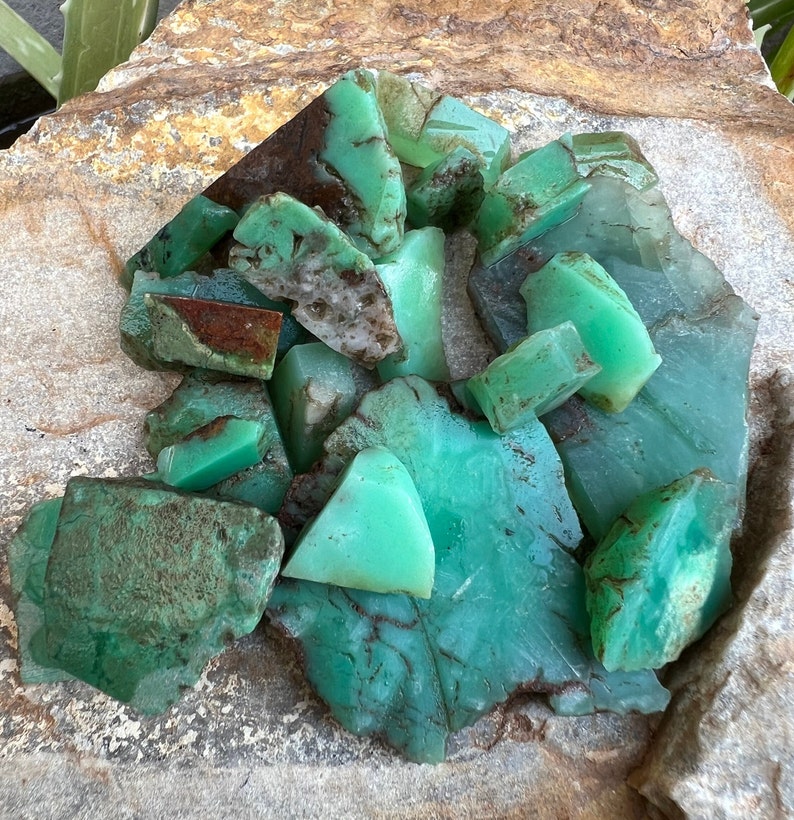 May include: A collection of green and brown chrysoprase gemstones, some with a rough, natural texture, others with a smooth, polished surface. The stones are arranged in a pile on a light brown, textured surface.