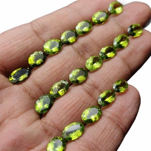 May include: A collection of oval-shaped green peridot gemstones, arranged in rows on a hand. The stones are faceted and have a translucent appearance.