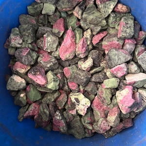 Könnte beinhalten: Eine Ansammlung von rohen, getrommelten Steinen mit einer Mischung aus leuchtend rosa und gedämpften grünen Mineralablagerungen. Die Steine sind unregelmäßig in Form und Größe und weisen eine raue, natürliche Textur im gesamten Haufen auf.