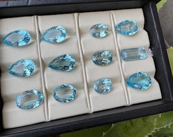 May include: A black tray filled with various light blue gemstones. The gemstones are in different shapes, including oval, pear, and rectangular cuts. The tray has a cream-colored interior with individual compartments for each stone.