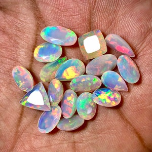 Pode incluir: Uma coleção de 14 opalas soltas, em forma de pêra e oval, em vários tons de branco, com flashes de arco-íris iridescentes. As opalas são exibidas em uma mão.