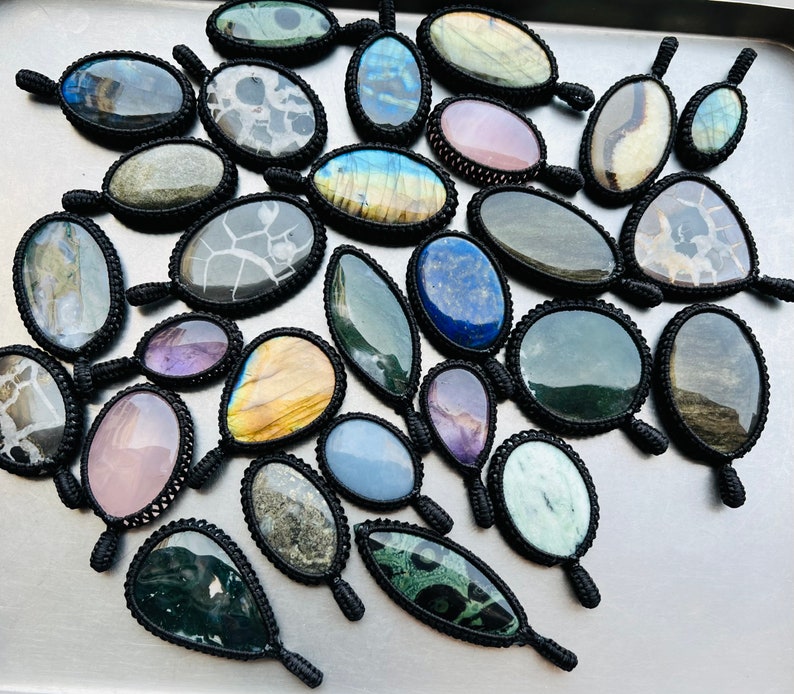 Peut inclure: Une collection de 20 pendentifs en pierres pr&eacute;cieuses uniques, chacun envelopp&eacute; d'un cordon en macram&eacute; noir. Les pierres sont de couleurs et de motifs vari&eacute;s, notamment le vert, le bleu, le violet, le rose et le brun.