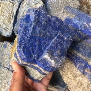 Pode incluir: Uma coleção de pedras de lápis-lazúli em bruto. As pedras são de cor azul profundo com veios brancos e dourados. A textura áspera das pedras é visível. A imagem mostra um close-up das pedras, destacando sua beleza natural.