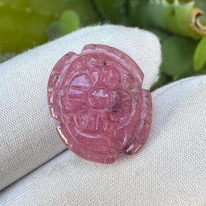 May include: A carved pink gemstone pendant with a floral design. The pendant is oval-shaped and features intricate details, with a central circular element surrounded by petal-like carvings. The gemstone has a translucent quality and is set against a white background.