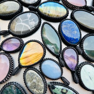 Peut inclure: Assortiment de pendentifs en pierres pr&eacute;cieuses de forme ovale avec cordon noir. Les pierres sont de diff&eacute;rentes couleurs et motifs, notamment vert, bleu, violet, rose et marron.