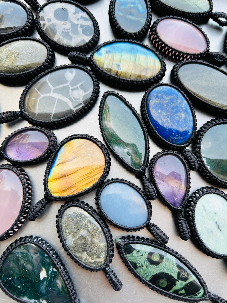 Peut inclure: Assortiment de pendentifs en pierres pr&eacute;cieuses de forme ovale avec cordon noir. Les pierres sont de diff&eacute;rentes couleurs et motifs, notamment vert, bleu, violet, rose et marron.