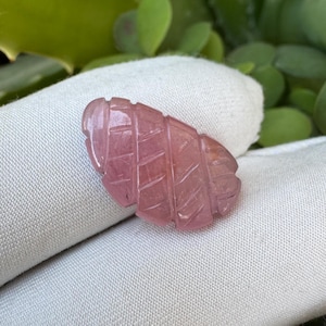 May include: A carved pink gemstone in the shape of a leaf. The gemstone has a textured surface with etched lines, and is set against a white cloth background. The gemstone is a light pink colour.