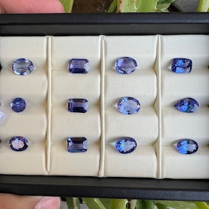 May include: A white tray with 12 compartments, each containing a different cut and color of blue gemstone. The gemstones are arranged in rows of three.