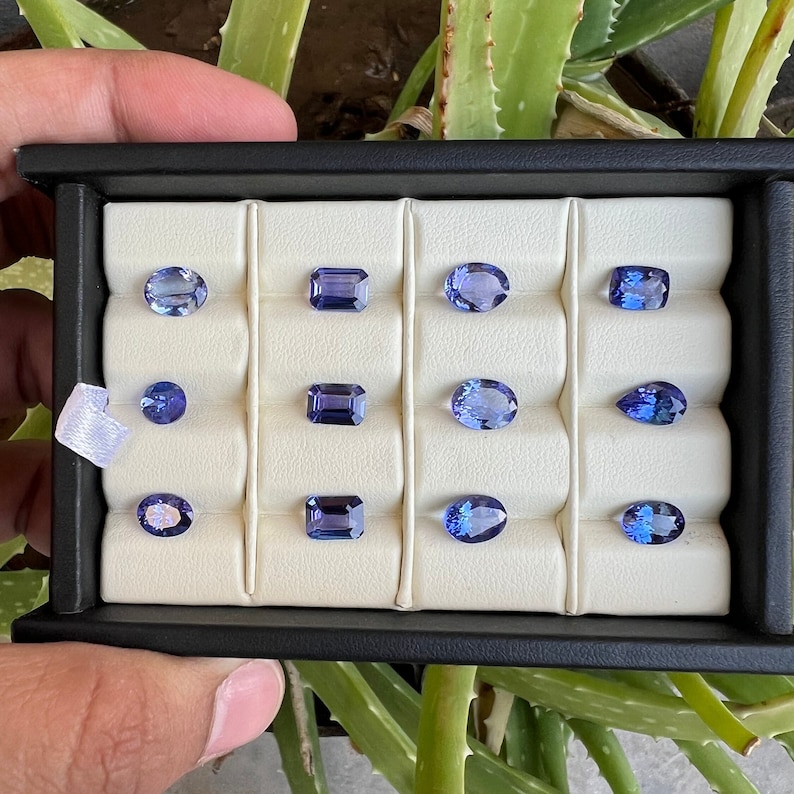 May include: A collection of twelve blue gemstones displayed in a white velvet-lined box. The gemstones are arranged in three rows of four, with each gemstone in its own individual compartment. The gemstones are various shapes, including oval, pear, and emerald cut.