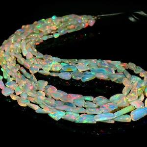 May include: Strands of iridescent opal beads. The beads are a mix of white, blue, green, and red, with a polished, smooth surface. The beads are strung together, creating a flowing, organic shape. The background is black.
