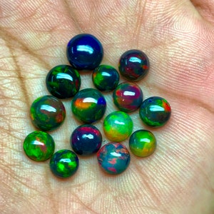 May include: A collection of 14 round black opal cabochons in various shades of green, red, and blue. The opals are displayed on a hand.