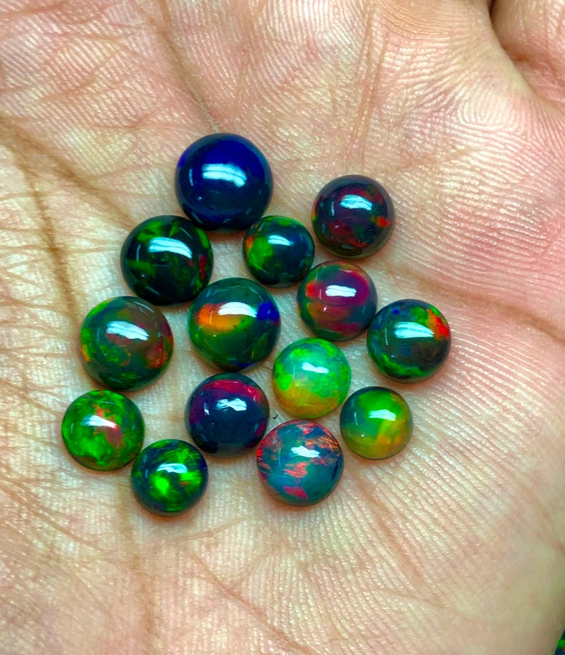 May include: A collection of 14 round black opal cabochons in various shades of green, red, and blue. The opals are displayed on a hand.