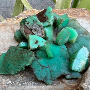 May include: A collection of green and brown chrysoprase gemstones. The stones are rough and have a natural, earthy look.
