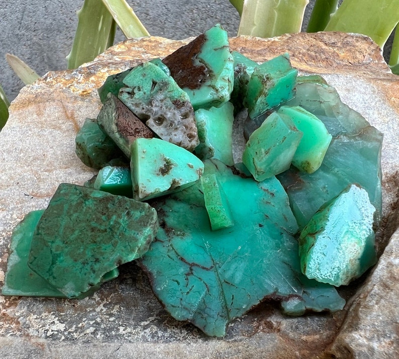 May include: A collection of green and brown chrysoprase gemstones. The stones are rough and have a natural, earthy look.