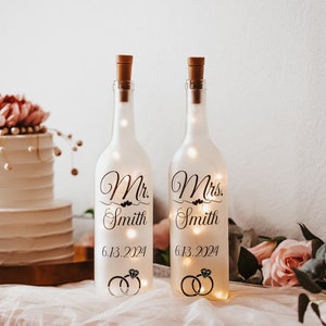 May include: Two illuminated glass bottles with cork stoppers, labeled "Mr. Smith" and "Mrs. Smith" with the date "6.13.2024" and ring graphics. A white wedding cake and floral arrangements are in the background.