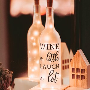 May include: A frosted glass wine bottle with a cork top and string lights inside. The bottle has a black decal that reads "Wine a little laugh a lot". The bottle is sitting on a stack of books with the words "home sweet farmhouse" printed on the top book.