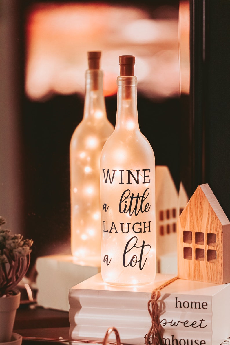 May include: A frosted glass wine bottle with a cork top and string lights inside. The bottle has a black decal that reads "Wine a little laugh a lot". The bottle is sitting on a stack of books with the words "home sweet farmhouse" printed on the top book.