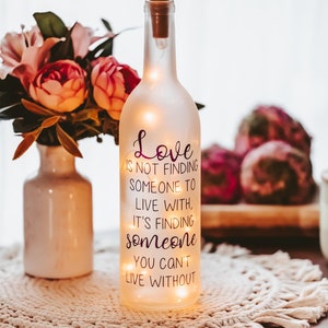 May include: A frosted glass bottle with a cork top and string lights inside. The bottle has a quote printed on it that reads: "Love is not finding someone to live with, it's finding someone you can't live without."