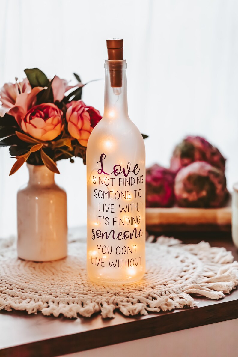 May include: A frosted glass bottle with a cork top and string lights inside. The bottle has a quote printed on it that reads: "Love is not finding someone to live with, it's finding someone you can't live without."