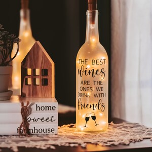 May include: A clear glass wine bottle with a cork top and string lights inside. The bottle has a black vinyl sticker with the text "The best wines are the ones we drink with friends" and a graphic of two wine glasses clinking.