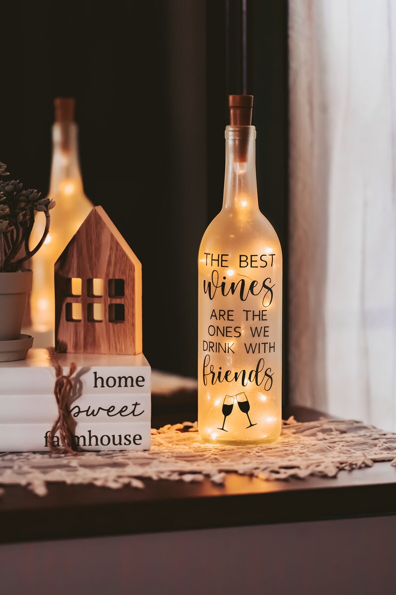 May include: A clear glass wine bottle with a cork top and string lights inside. The bottle has a black vinyl sticker with the text "The best wines are the ones we drink with friends" and a graphic of two wine glasses clinking.
