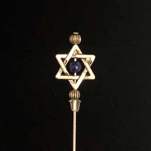 Peut inclure: Une épingle à cravate dorée représentant une étoile de David avec une pierre précieuse bleue au centre. L'épingle est ornée d'une boule décorative en haut et en bas.