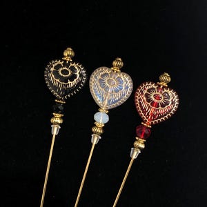 Puede incluir: Tres alfileres de vidrio en forma de corazón dorado con intrincados diseños florales. Los corazones son negros, blancos y rojos. Cada alfiler tiene una cuenta dorada y una cuenta de cristal transparente.