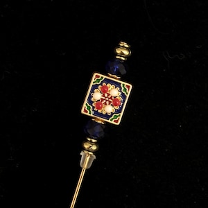 May include: A decorative hat pin with a square, enameled top featuring a floral design in red, white, and green on a blue background. The pin has gold-colored accents and a gold-toned pin.