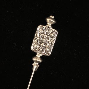 May include: A silver-toned hat pin with a rectangular, ornate head featuring a floral design. The pin has a decorative top and a long, slender needle. The background is a dark, solid color.