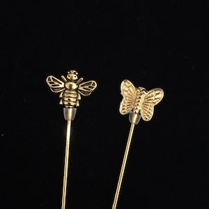 May include: Two gold-toned pins with decorative tops. One pin features a bee design, the other a butterfly design.