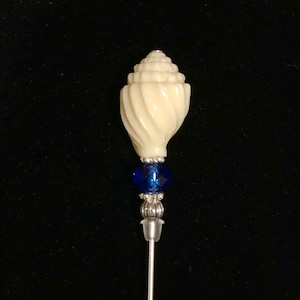 May include: A white seashell-shaped pin with a blue crystal bead and a silver metal base. The pin is attached to a long, thin silver metal rod.