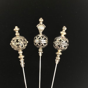 Puede incluir: Tres alfileres decorativos plateados con tapas de metal ornamentadas. Las tapas son redondas y tienen diseños intrincados. Los alfileres son largos y delgados.