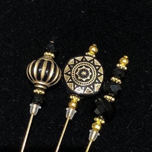 May include: Three gold and black decorative hat pins with black beads. The pins feature a striped sphere, a geometric design, and a string of black beads.