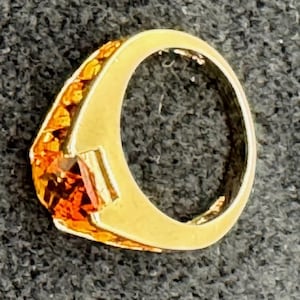 Könnte beinhalten: Ein goldener Ring mit einem orangefarbenen Edelstein. Der Ring hat einen rechteckigen Edelstein und ist von kleineren orangen Steinen umgeben. Das Band hat eine goldene Farbe.
