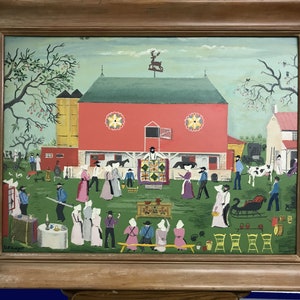 Pode incluir: Pintura emoldurada de arte popular retratando uma cena rural com um celeiro vermelho, figuras em trajes de época e uma exposição de colchas. A obra apresenta um campo verde, um silo amarelo e uma cata-vento de veado.