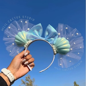 May include: A pair of light blue and white seashell-shaped Minnie Mouse ears with a white tulle bow and pearl accents. The ears are on a white headband. The text "Lil Cactus Studio" is written on the headband.