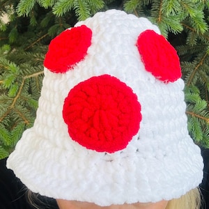 May include: A white, crocheted bucket hat with three large, red, circular accents. The hat is made of a soft, textured yarn. The hat is shown against a backdrop of green pine needles.