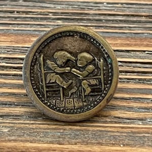 May include: A round, antique brass button with a detailed relief of two figures seated at a table. The figures appear to be children, engaged in an activity. The button has a decorative border and is set against a wooden surface.