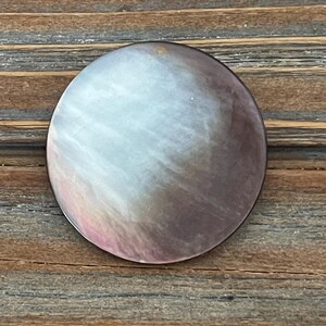 May include: A circular button featuring a smooth, iridescent surface with a gradient of colors transitioning from a soft, pale white and light pink on one side to a deep, dark brown and charcoal gray on the other side, displaying a natural luster.
