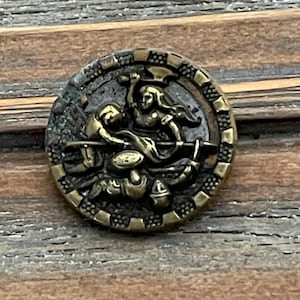 May include: A round, antique brass button with a detailed relief scene. The button features a central design of figures in a struggle, surrounded by a decorative border. The button is set against a wooden background.