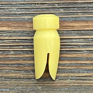 May include: A pale yellow plastic tooth-shaped object with a split base. The object has a cylindrical top and a wider, tapered body. The background is a wooden surface with horizontal grain.