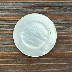 May include: A round, iridescent, mother-of-pearl button with a smaller, raised circle in the center. The button has a pearlescent sheen and is set against a dark wood grain background. This button is suitable for sewing projects.