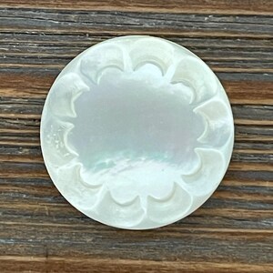 May include: A round, iridescent mother-of-pearl button with a scalloped edge. The button has a smooth, reflective surface and is set against a dark wood grain background. The button's design is simple yet elegant.