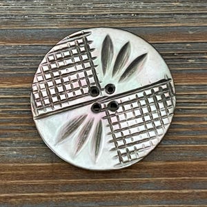 May include: A round, iridescent mother-of-pearl button featuring a four-hole center. The surface is divided into four quadrants, alternating between a textured grid pattern and a smooth section with three carved, leaf-like petal shapes.