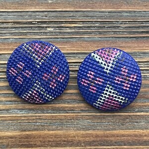 May include: Two round blue fabric earrings with a textured surface. The earrings have a pink and white pattern. The earrings are displayed on a wooden surface.