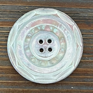 May include: A round, four-hole button with an iridescent, pearlescent finish. The button features a recessed center and a raised, segmented outer rim with a subtle, light-reflecting sheen that shifts between pale pink, blue, and white tones.