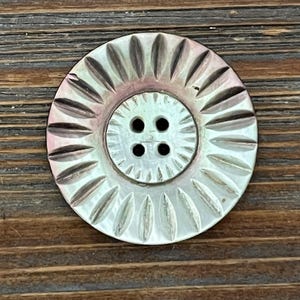 May include: A round, iridescent mother-of-pearl button featuring a central recessed area with four thread holes. The button has a decorative sunburst pattern of carved, radiating grooves extending from the center to the outer edge.