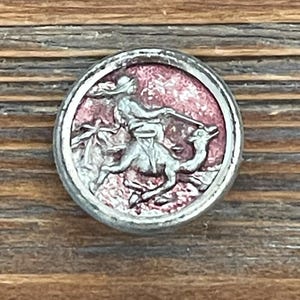 May include: A round, silver-toned button with a raised relief design. The central image depicts a figure on horseback, possibly a knight or warrior, riding a camel-like animal. The background is a mottled pink color.