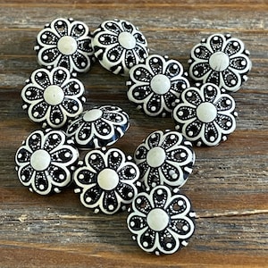 May include: Ten white and black floral buttons with a raised design. The buttons are arranged in a semi-circle on a wooden surface.